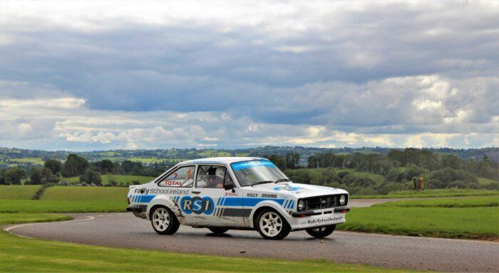 Rally car driving on a winding road with scenic countryside in the background under a cloudy sky.