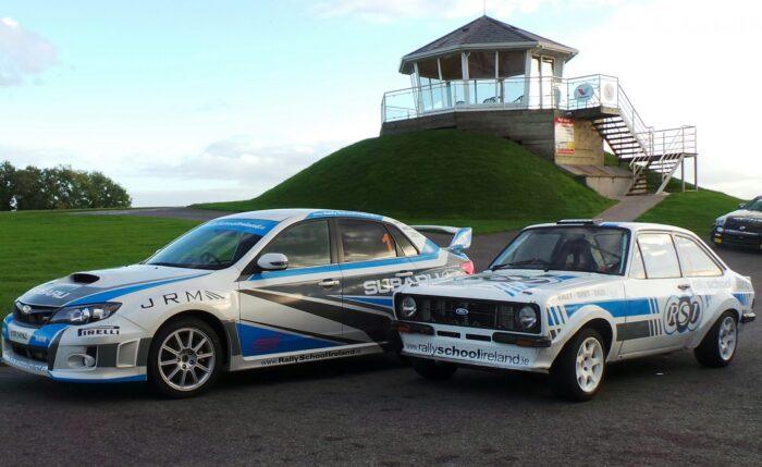 Rally cars at a track with a pavilion in the background, showcasing Subaru and Ford models.