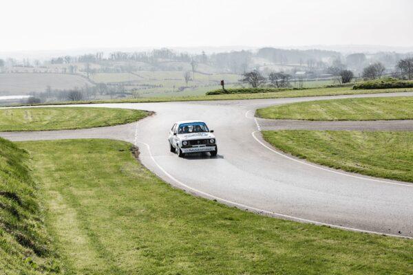 Vintage rally car navigating a scenic countryside track with rolling hills in the background.