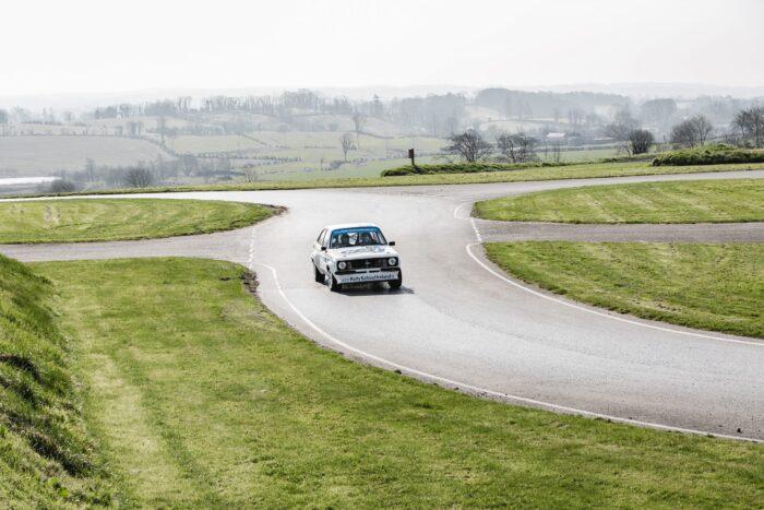 Vintage rally car navigating a scenic countryside track with rolling hills in the background.