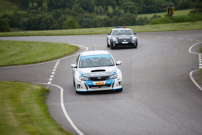 Race car leading on a winding road followed by a police car in a green, scenic landscape.