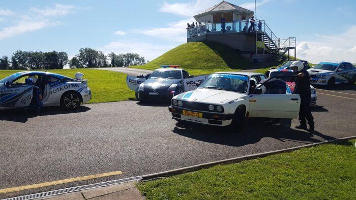 Race cars and police cars on a track, drivers and instructors preparing, a scenic view with a hill and building in the background.