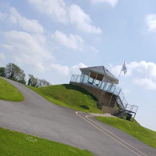 Hilltop building with glass railings and a flag under a blue sky, surrounded by green grass and a winding road.