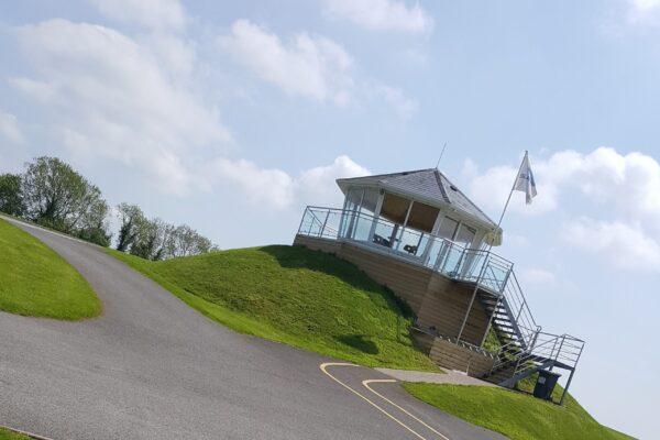 Hilltop building with glass railings and a flag under a blue sky, surrounded by green grass and a winding road.