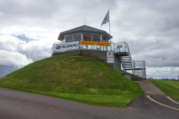 Tower on grassy hill with Subaru and Pirelli banners under cloudy sky. Metal stairway leads to the entrance.