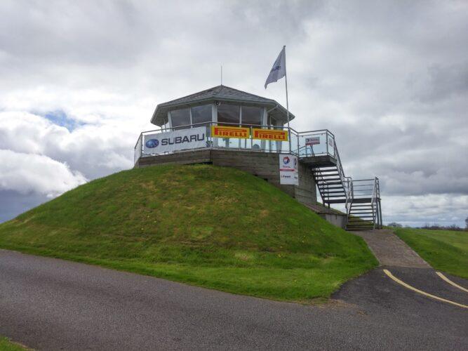 Tower on grassy hill with Subaru and Pirelli banners under cloudy sky. Metal stairway leads to the entrance.