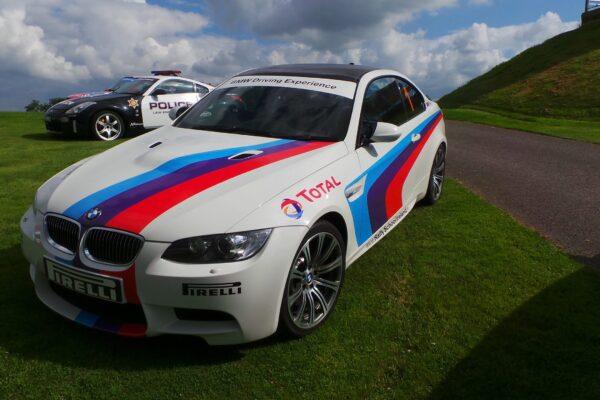 White BMW sports car with red and blue racing stripes on green grass, featuring a BMW Driving Experience banner.