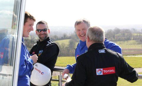 Group of race car drivers in blue and black suits chatting and smiling outdoors on a sunny day.