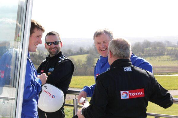 Group of race car drivers in blue and black suits chatting and smiling outdoors on a sunny day.