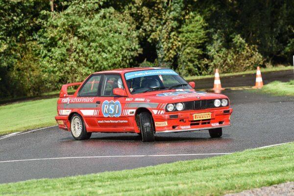 Red rally car from Rally School Ireland drifting on a wet track with trees in the background.