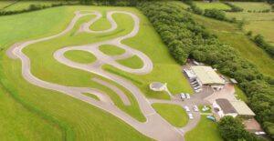 Aerial view of a winding outdoor track on lush green landscape with nearby buildings and parked cars.