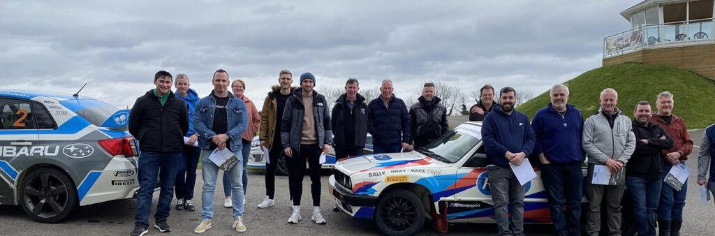 Group of people standing next to rally cars at a motorsport event on a cloudy day.