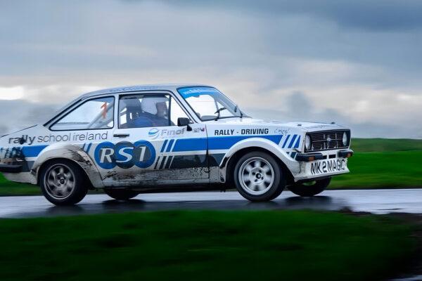 Vintage rally car on wet road, showcasing speed and control, with lush green landscape blurred in the background.