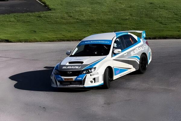 White and blue Subaru rally car drifting on a race track with green grass background.