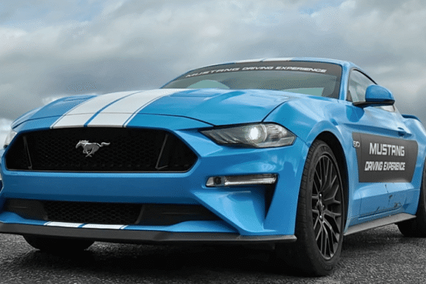 Blue Ford Mustang with racing stripes parked on a road, promoting a driving experience, under cloudy skies.